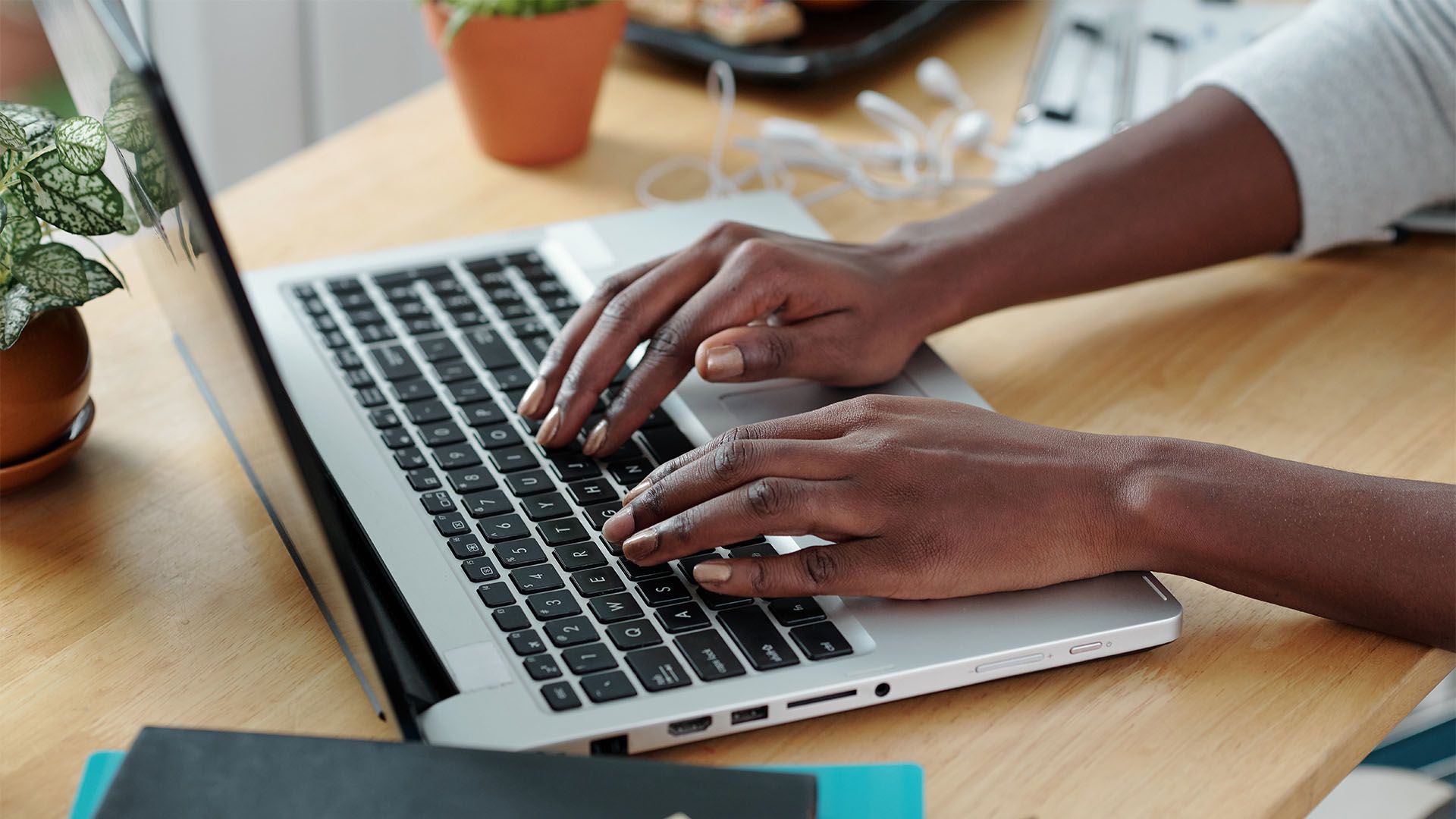 Pessoa digitando em um laptop em uma mesa de madeira com uma planta e fones de ouvido por perto.