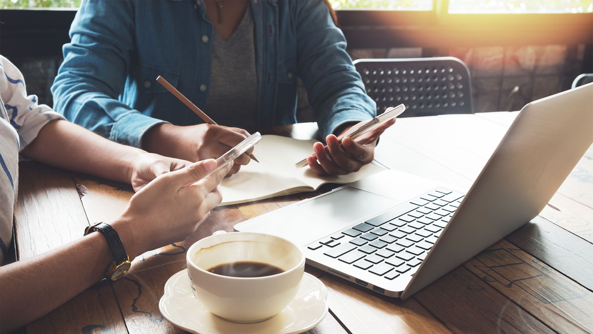 Duas pessoas sentadas em uma mesa de madeira, com laptops, celulares, café e cadernos, trabalhando juntas.