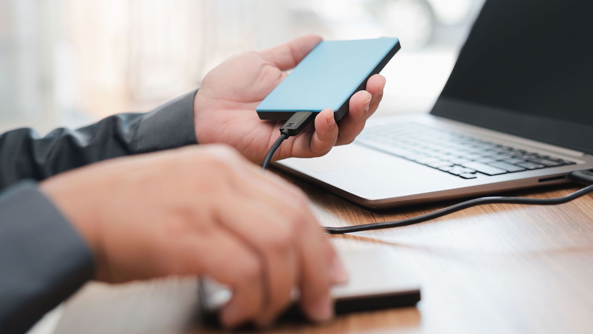 Pessoa conectando um disco rígido externo azul a um laptop com um cabo USB. Outro disco rígido está sobre a mesa.