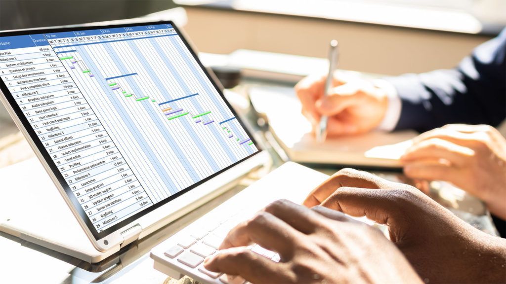 Mãos digitando em um laptop com um cronograma de projeto visível. Alguém fazendo anotações em um caderno.