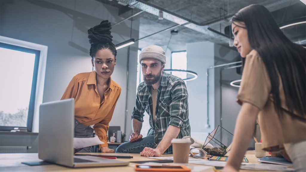Três pessoas se reúnem em torno de um laptop em um escritório moderno, revisando documentos.