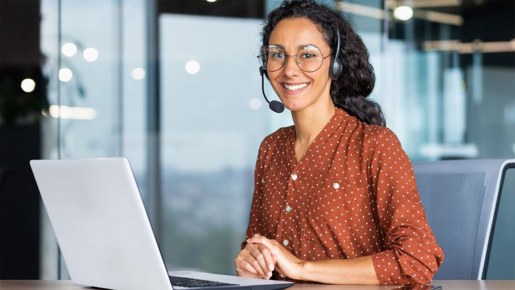 Mulher usando fones de ouvido e óculos sorri enquanto está sentada em frente a um laptop em um escritório.