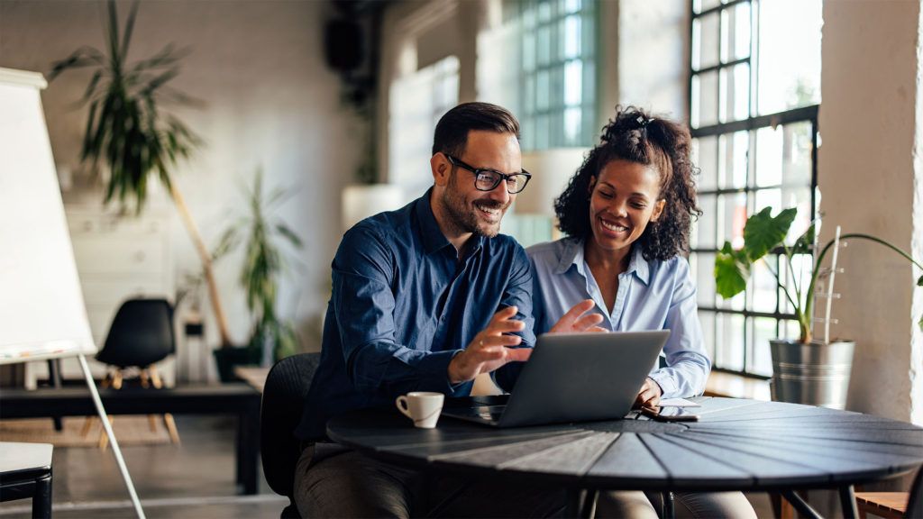 Homem e mulher sorrindo para um laptop, sentados à mesa em um escritório bem iluminado.