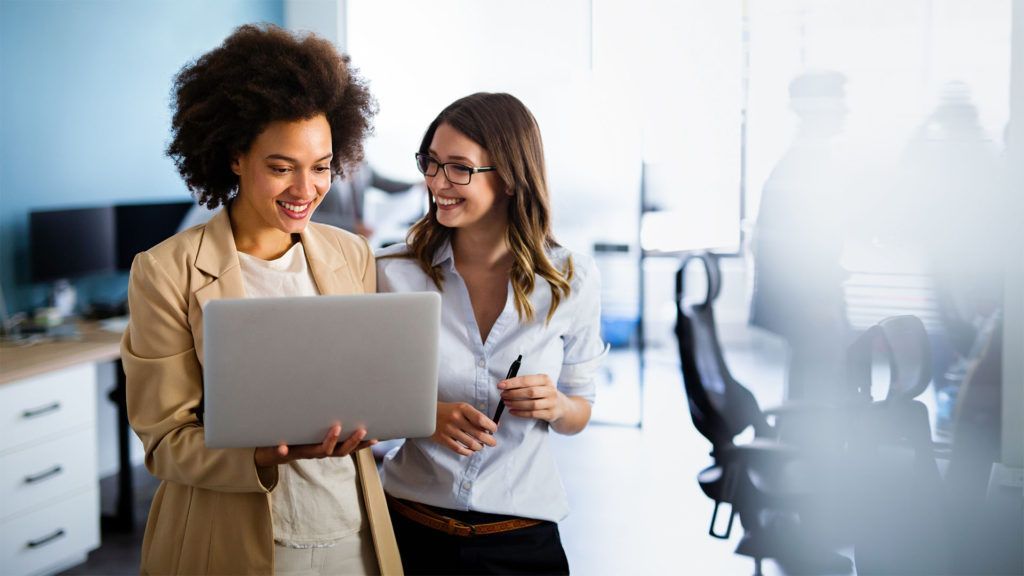 Duas mulheres sorrindo, olhando para um laptop em um escritório moderno, sob a luz do sol, com pessoas ao fundo.