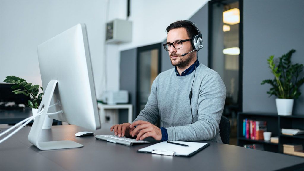 Homem de óculos e fone de ouvido digita em um computador em um ambiente de escritório.
