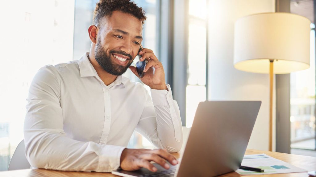 Homem sorrindo, falando ao telefone, trabalhando em um laptop em uma mesa perto de uma janela.