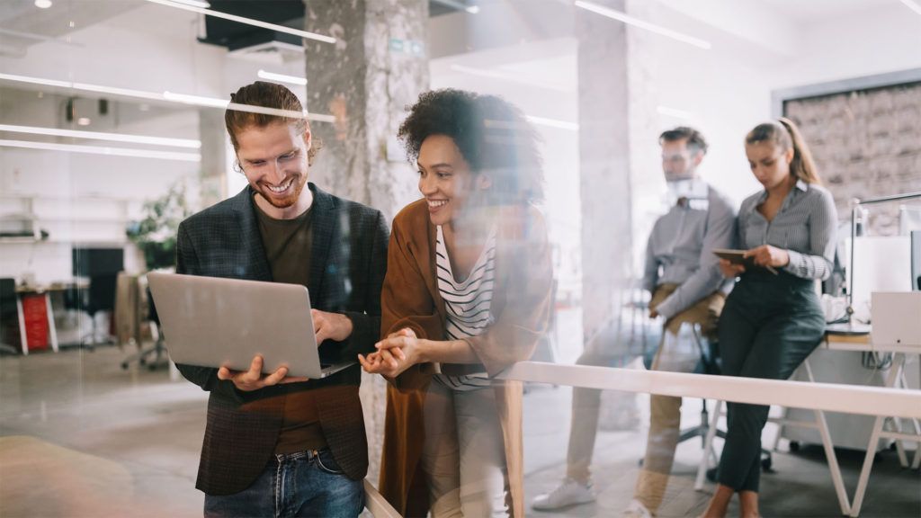 Dois colegas, um homem e uma mulher, sorrindo enquanto olham para um laptop em um ambiente de escritório.