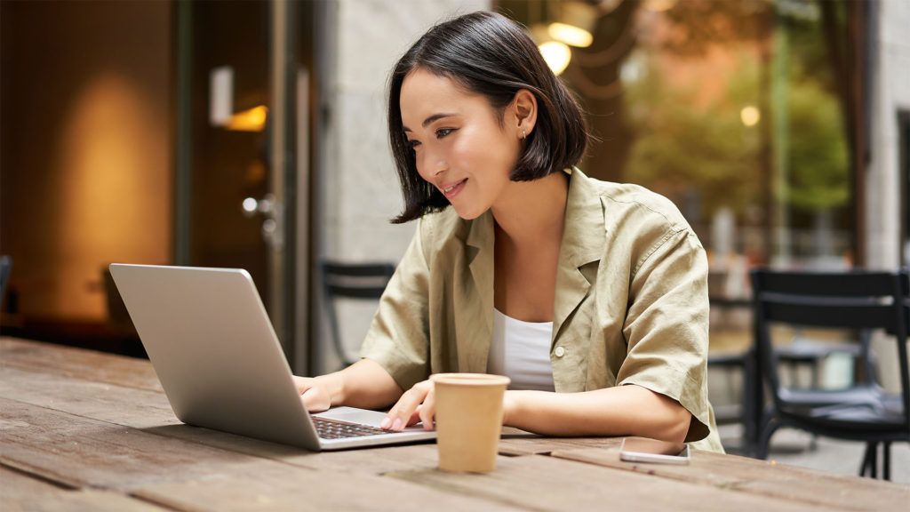 Mulher usando um laptop em uma mesa de café, sorrindo. Xícara de café à sua frente. Ambiente externo.