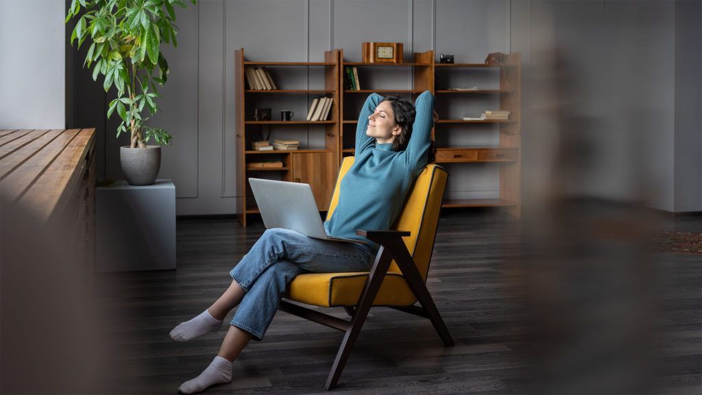 Mulher relaxando em uma cadeira amarela com um laptop, mãos atrás da cabeça, olhos fechados em seu escritório em casa.