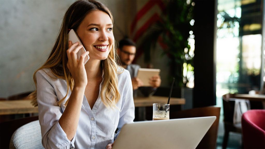 Mulher em um café, sorrindo enquanto fala ao telefone e usa um laptop; outro cliente desfocado ao fundo.