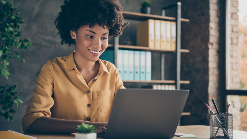 Mulher sorridente digitando em um laptop em sua mesa, em um ambiente de escritório moderno.
