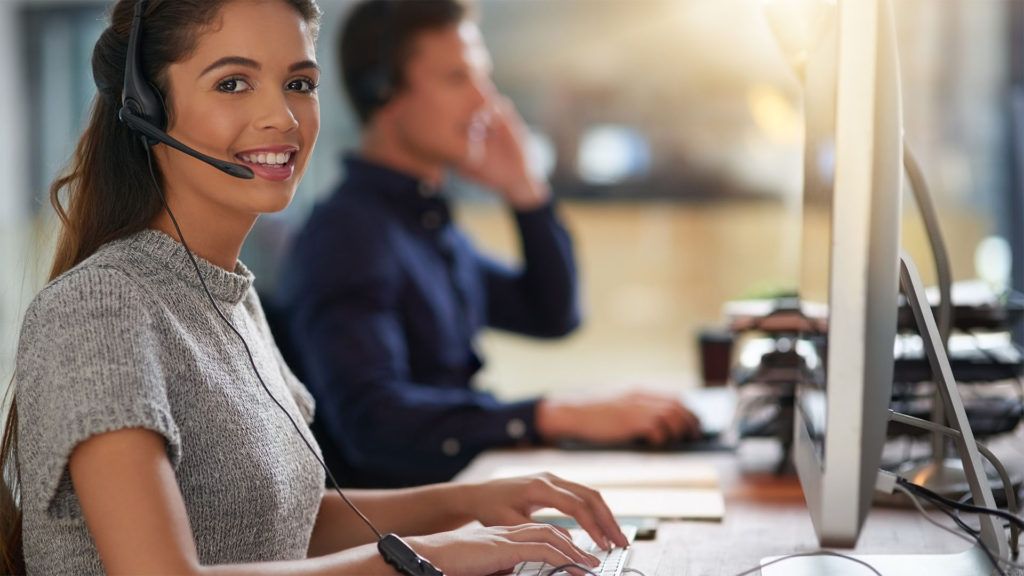 Mulher com fone de ouvido, sorrindo, digitando em um computador em um escritório bem iluminado; colega de trabalho ao fundo em uma chamada.