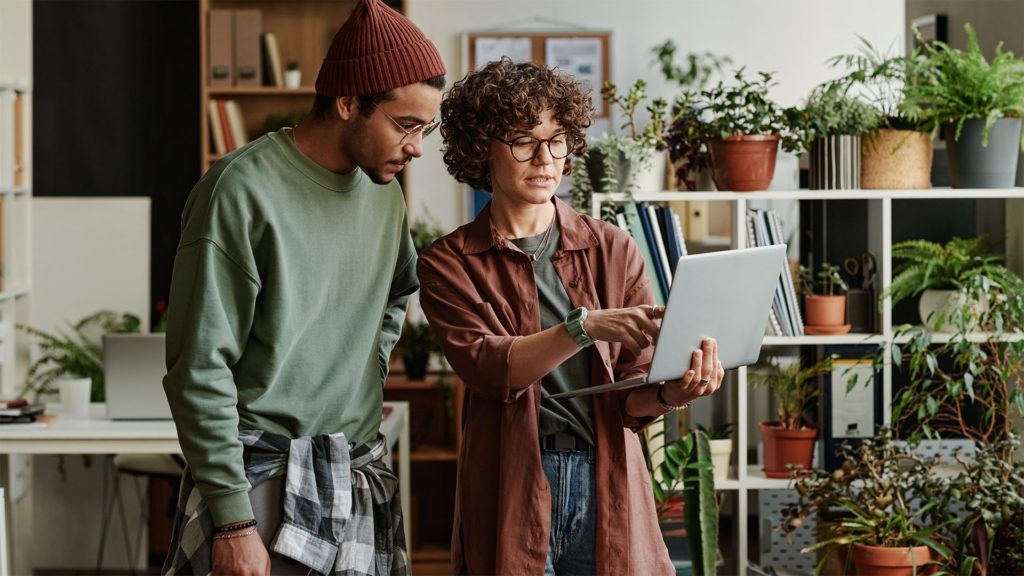 Duas pessoas olhando para um laptop em um escritório cheio de plantas. Uma delas aponta para a tela.