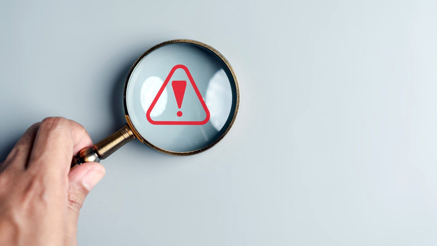 Mão segurando lupa sobre símbolo de alerta, representando gestão de riscos em TI e prevenção de falh