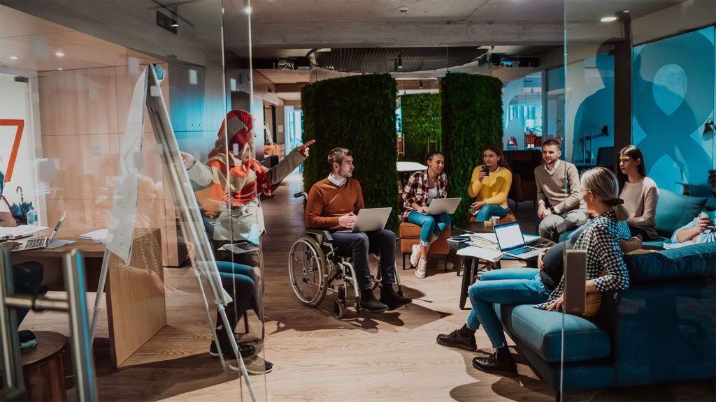 Um grupo de pessoas em um escritório assiste a uma apresentação. Entre elas, está uma pessoa em cadeira de rodas.