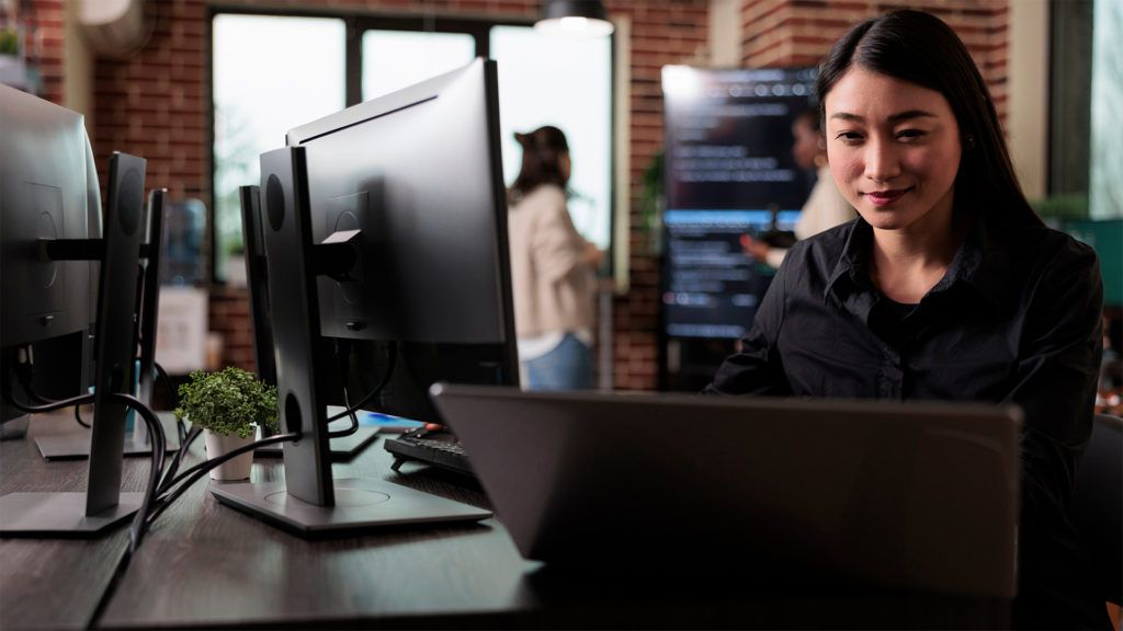 Mulher sentada à mesa, usando laptop e monitor em um ambiente de escritório, sorrindo para a câmera.