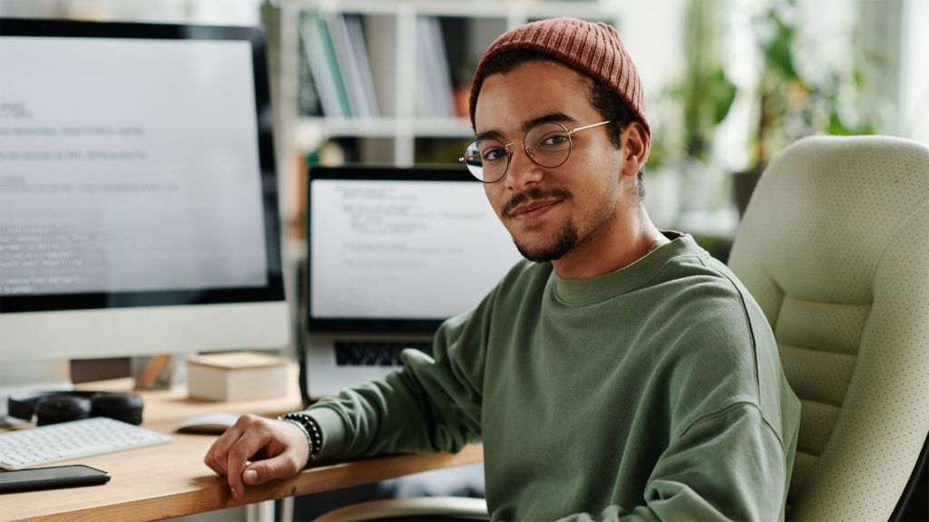 Homem de óculos e gorro sorri para a câmera enquanto está sentado em uma mesa com duas telas de computador.