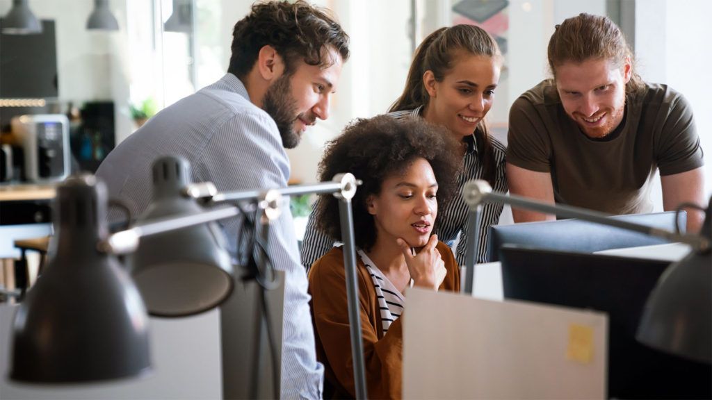 Quatro colegas de trabalho olhando para a tela do computador em um escritório, conversando sobre trabalho com sorrisos.