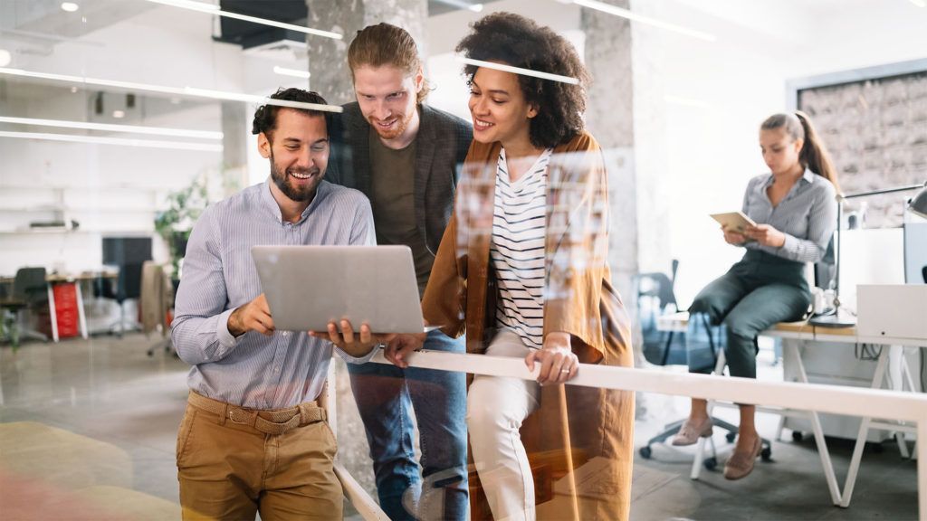 Três pessoas no escritório olhando para laptops, sorrindo. Uma mulher se apoia no corrimão; outra mulher usa um tablet.