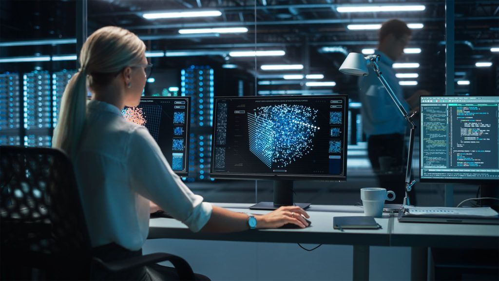 Mulher em frente a um computador com leitor de impressões digitais 3D; homem passa por perto na sala de servidores.