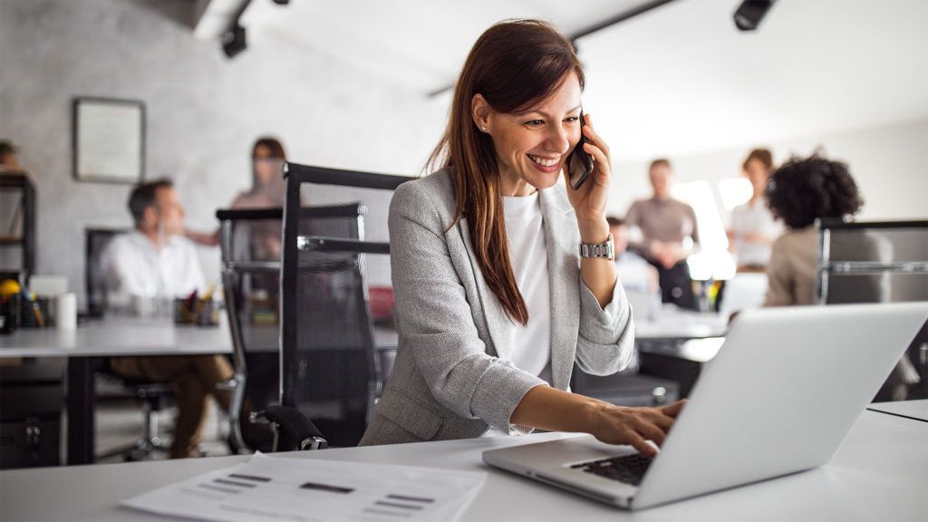 Mulher sorrindo enquanto fala ao telefone e usa um laptop em um escritório moderno com colegas de trabalho.