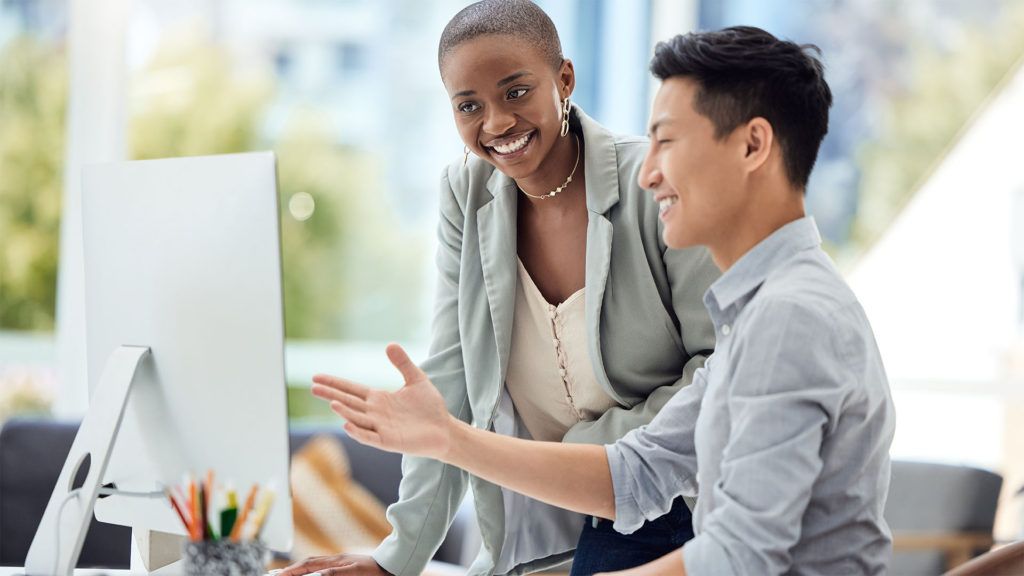 Uma mulher e um homem olham para a tela do computador, sorrindo. A mulher aponta, o homem gesticula com a mão no escritório.