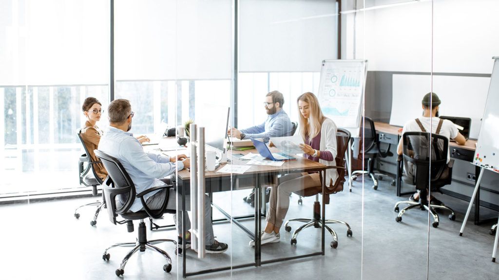 Reunião de escritório: Pessoas sentadas à mesa com laptops e documentos. Uma pessoa ao fundo trabalha em sua mesa. A luz do sol entra pela janela.