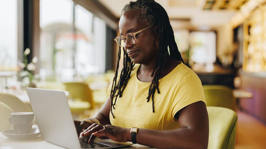 Mulher de camisa amarela e óculos, digitando em um laptop em um café.