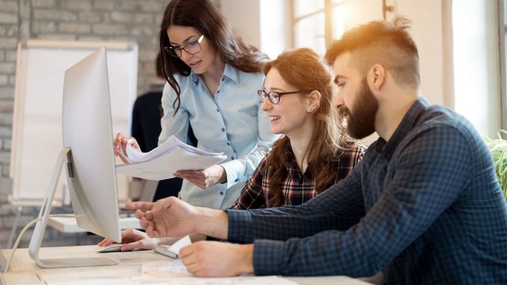 Três colegas de trabalho revisando documentos e um monitor de computador em um ambiente de escritório.