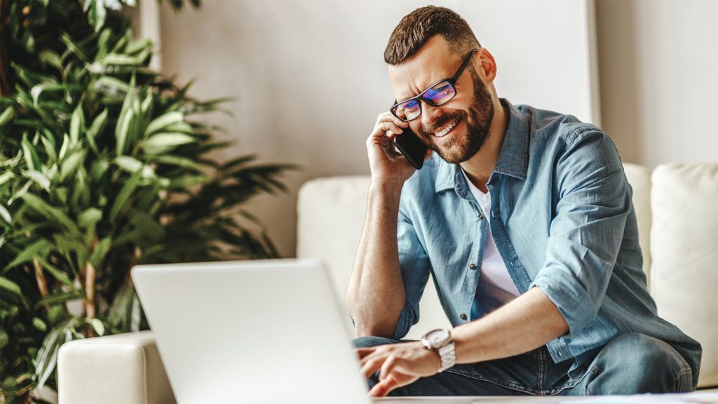 Homem sorrindo enquanto fala ao telefone, usando um laptop, sentado em um sofá perto de uma planta.
