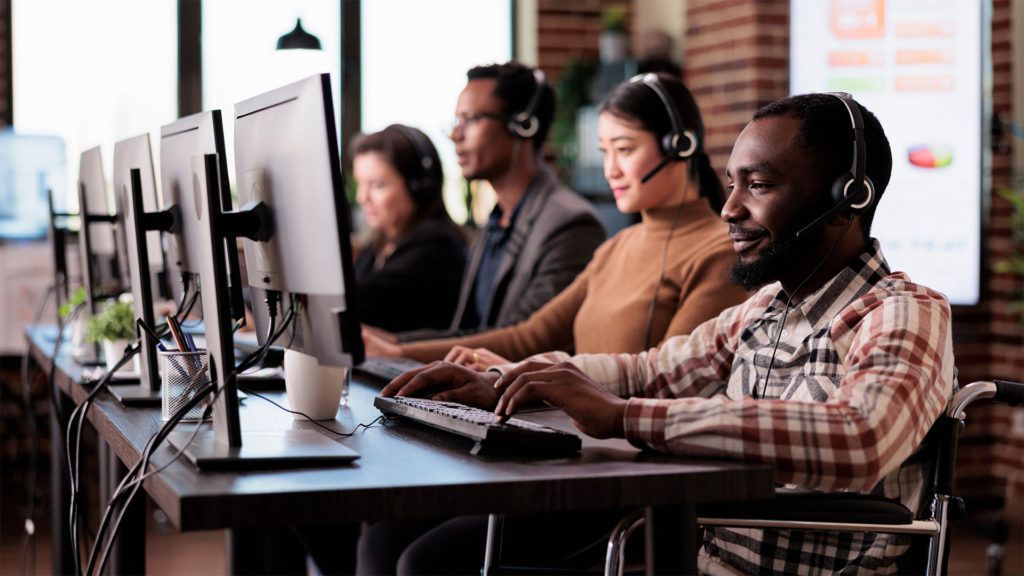 Pessoas com fones de ouvido trabalhando em computadores em um ambiente de escritório; uma pessoa usa cadeira de rodas.