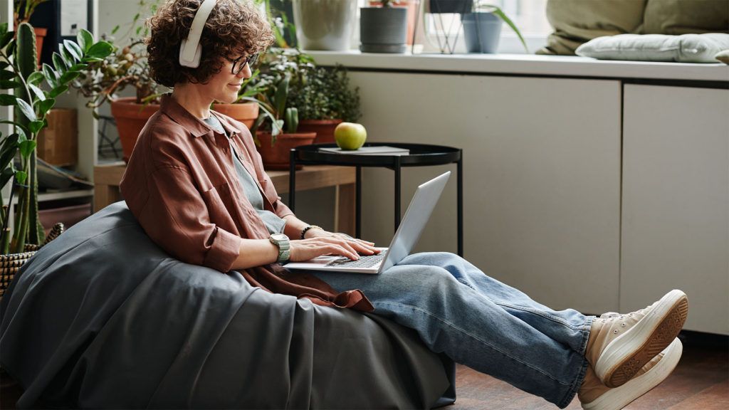 Mulher usando fones de ouvido e um laptop, sentada em uma poltrona tipo puff em uma sala com plantas.