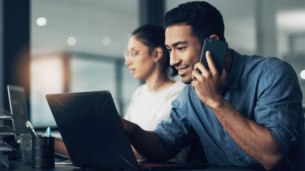 Homem ao telefone, olhando para um laptop em um ambiente de escritório. Uma mulher trabalha em um computador ao fundo.
