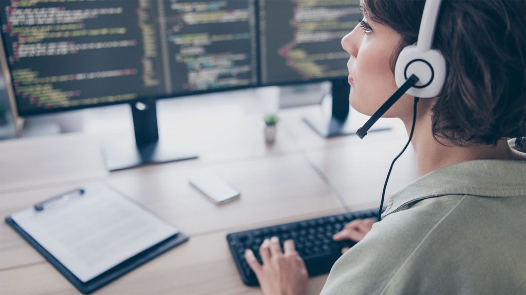 Mulher usando fones de ouvido, digitando em um teclado e olhando para telas de computador com código. Ambiente de escritório.