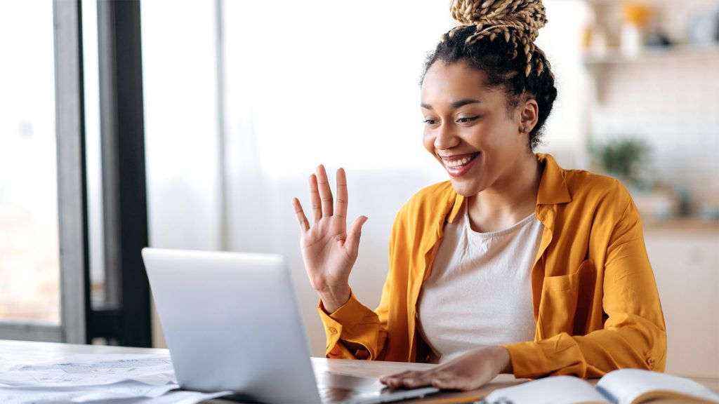 Mulher com tranças onduladas em frente à tela do laptop, sorrindo. Vestindo camisa laranja, em ambiente interno.