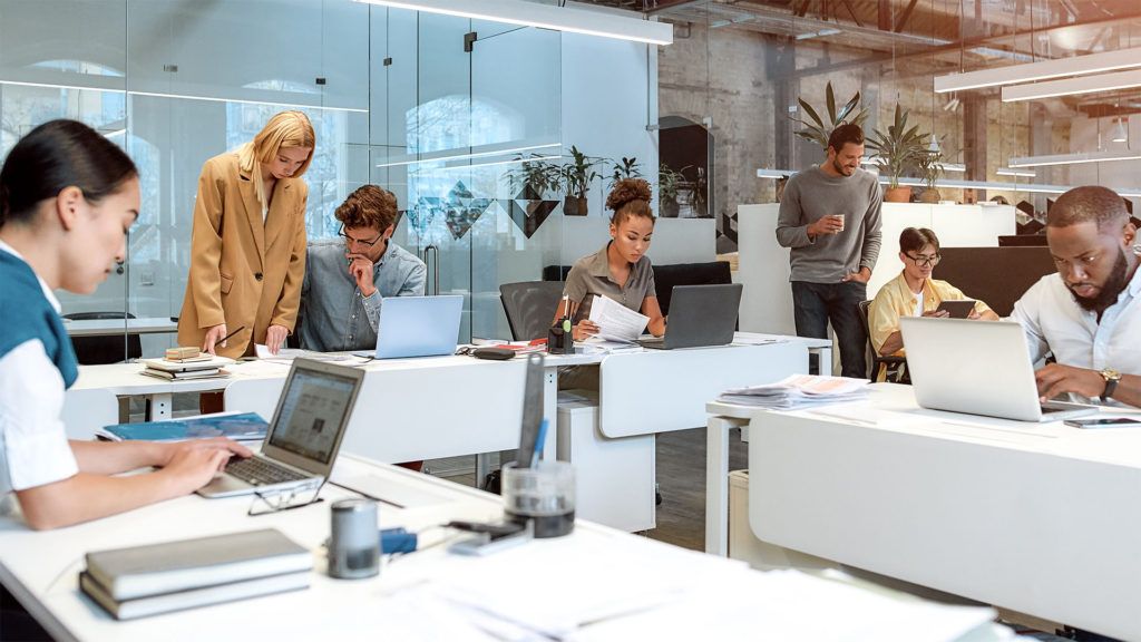 Pessoas trabalhando em mesas em um escritório moderno com laptops e papéis; ambiente colaborativo.