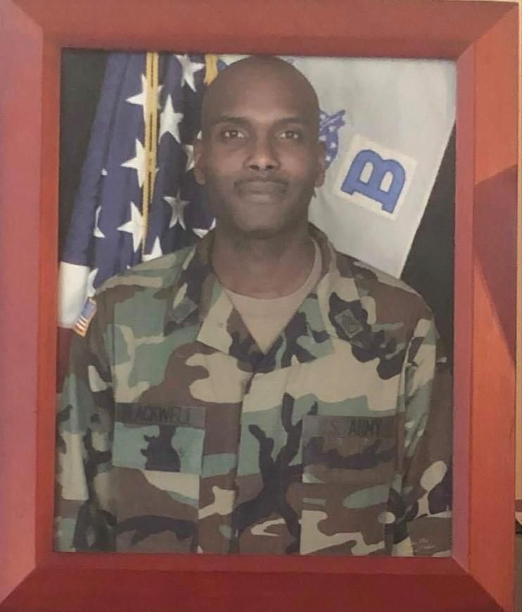 Man in military uniform, framed, with US flag and military banner in background.
