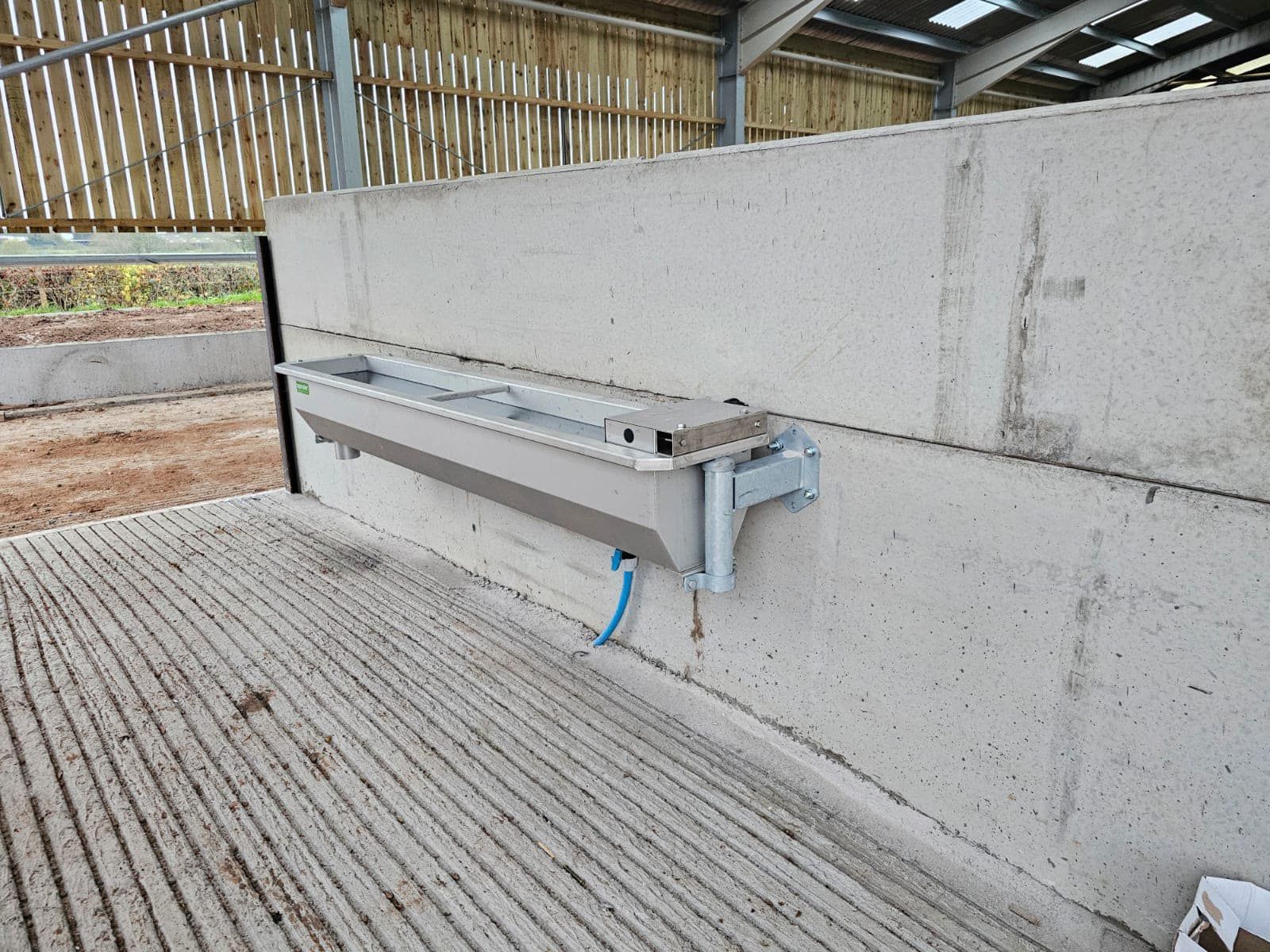 A metal livestock water trough mounted on a concrete wall inside an agricultural barn.