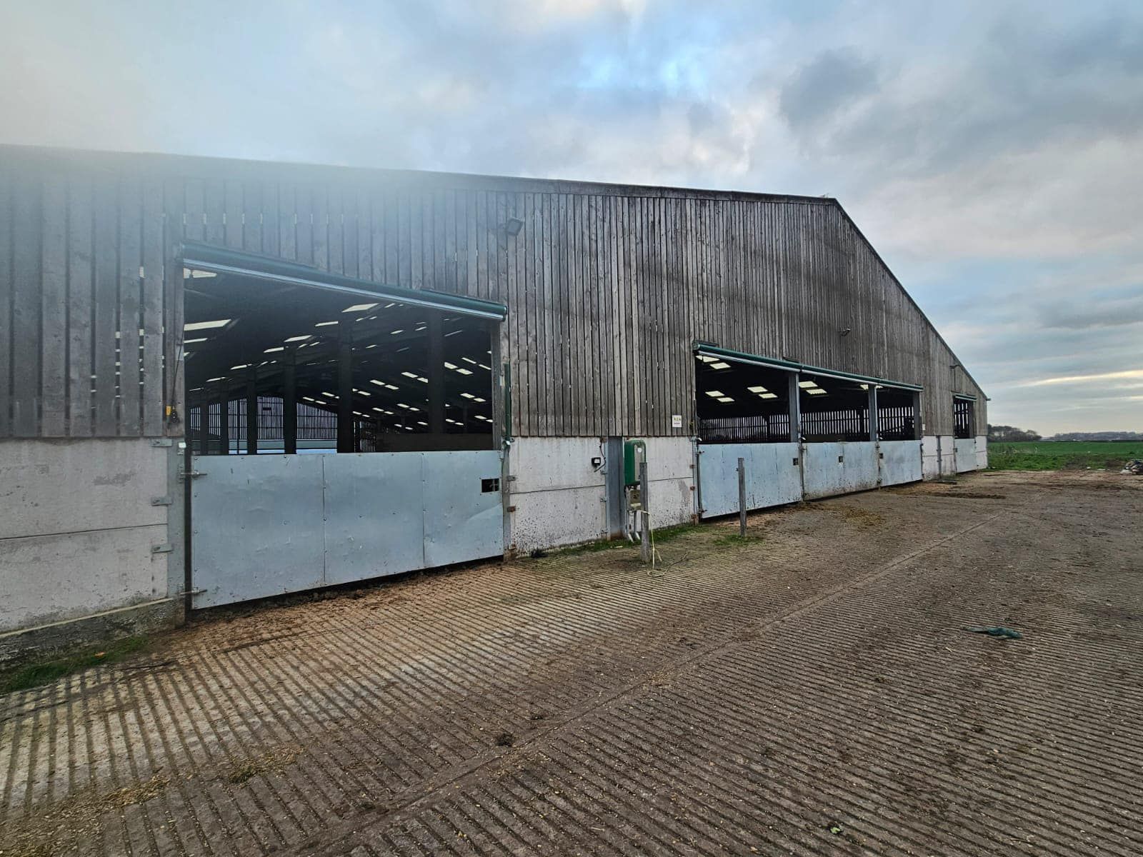 A large barn with weathered wooden siding, open bays, and a concrete-paved yard under a cloudy sky.