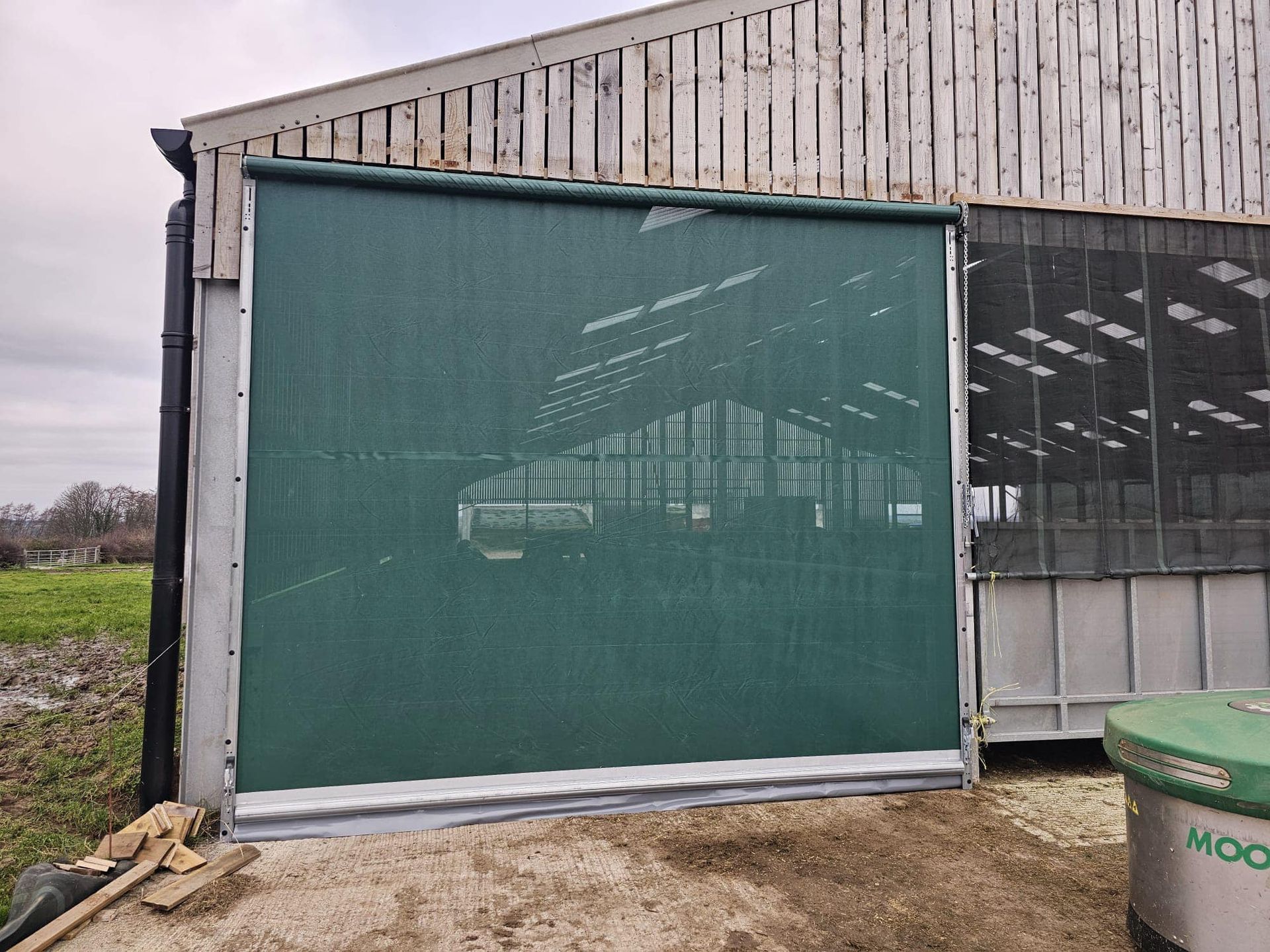 A green mesh curtain screen installed on the side of a farm building with wooden siding and a gravel ground area.