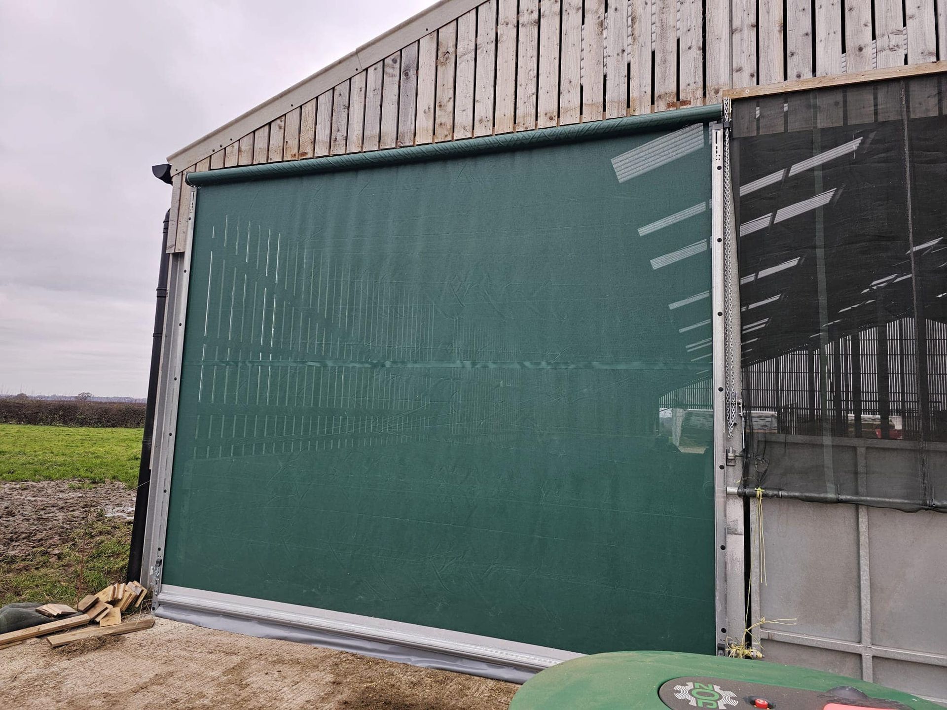 A large green roll-down mesh curtain installed on the side of a farm building with wooden siding.