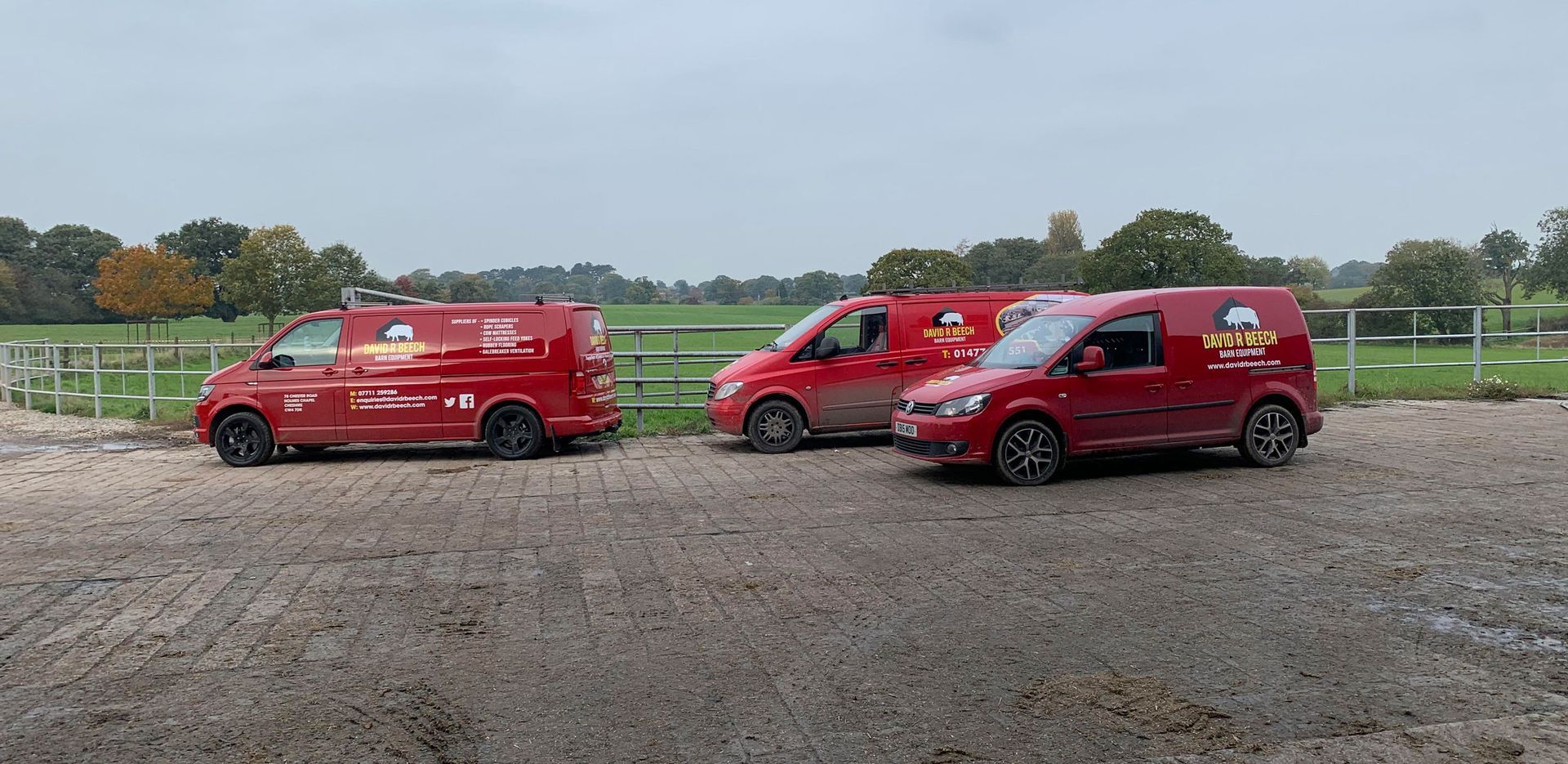 Three red work vans parked on a dirt lot in front of a grassy field.