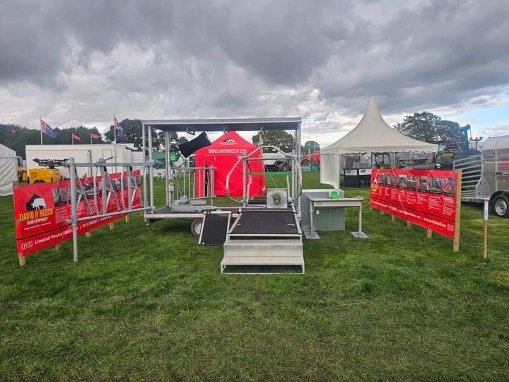 A trade show booth featuring a mobile cattle handling unit, a red tent, a white gazebo, and red signage on a grassy field.