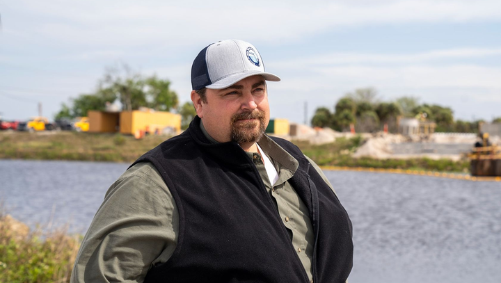 Ben Butler standing by a body of water, wearing a black vest and baseball cap, with trees and buildings in the background