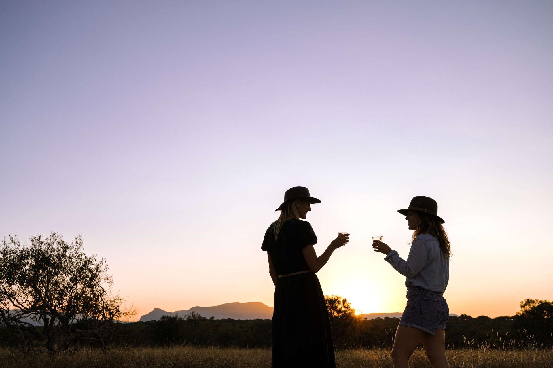 Two women are standing in a field drinking wine at sunset.
