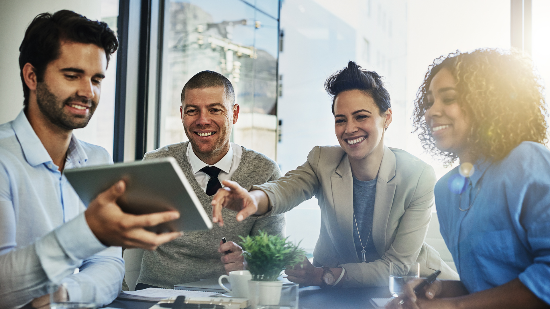 Four colleagues gathered in a bright office, smiling while looking at a tablet held by one person.
