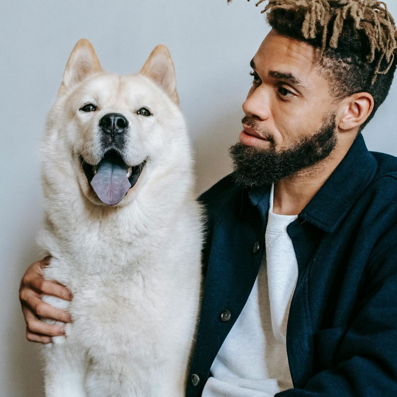 Man with a beard and dreadlocks smiles, next to a large white dog with a lolling tongue, both indoors.