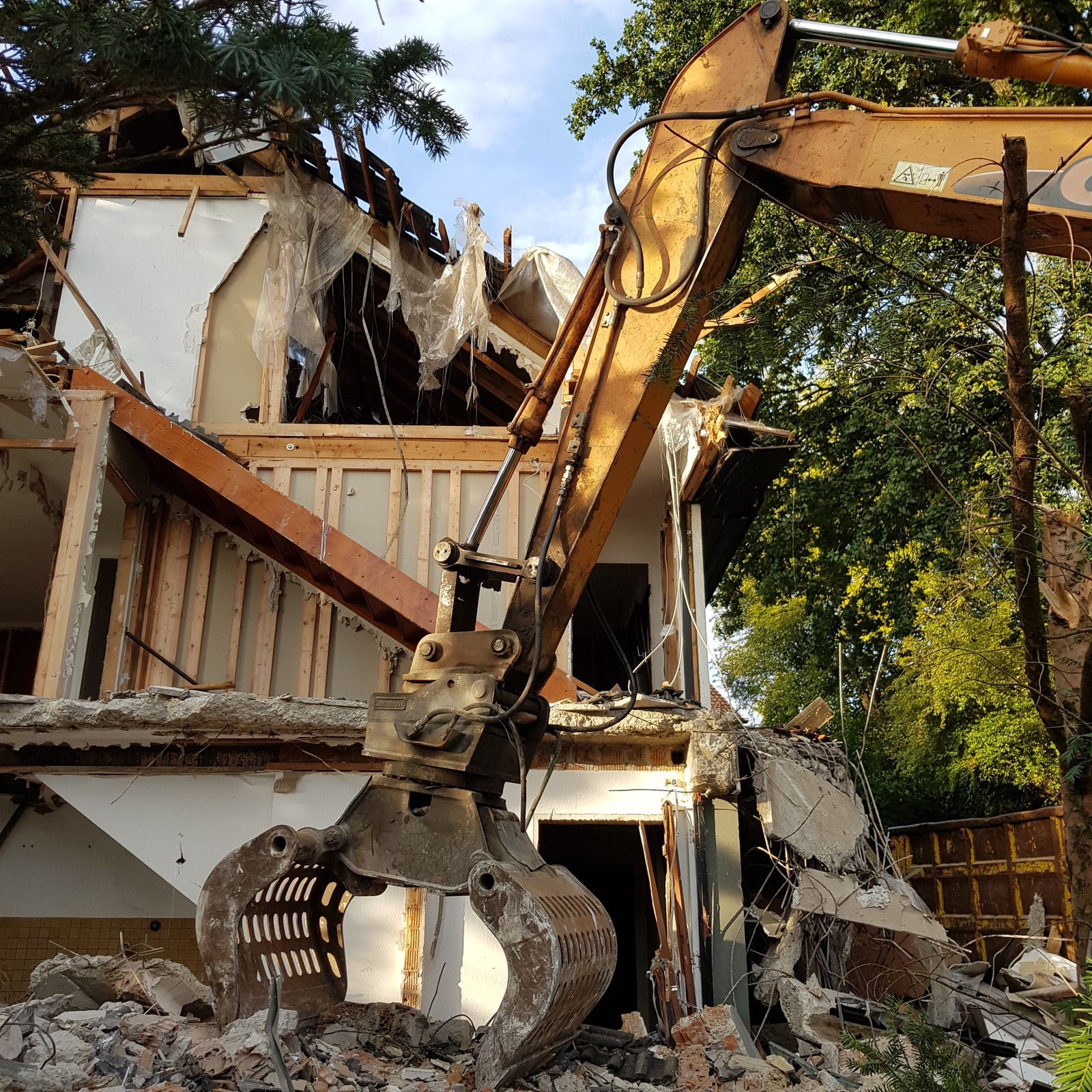 Excavator demolishing a two-story house; debris piles below.