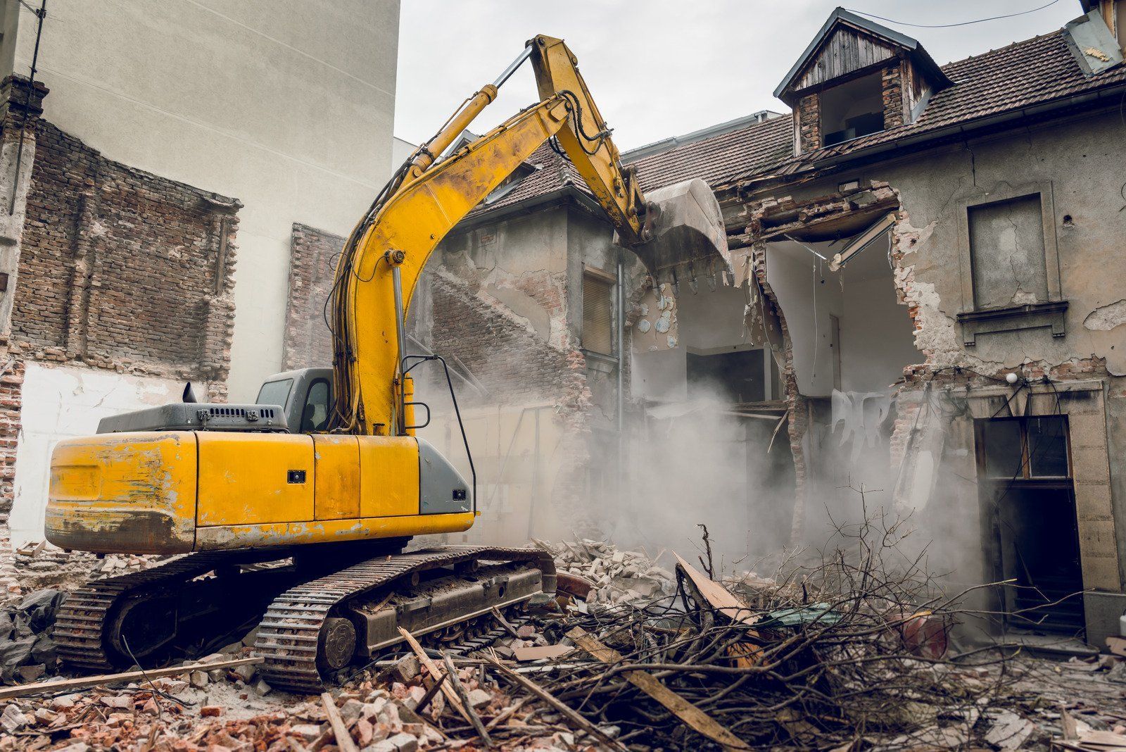 Yellow excavator demolishing a brick building, creating dust. Outdoor scene, overcast sky.