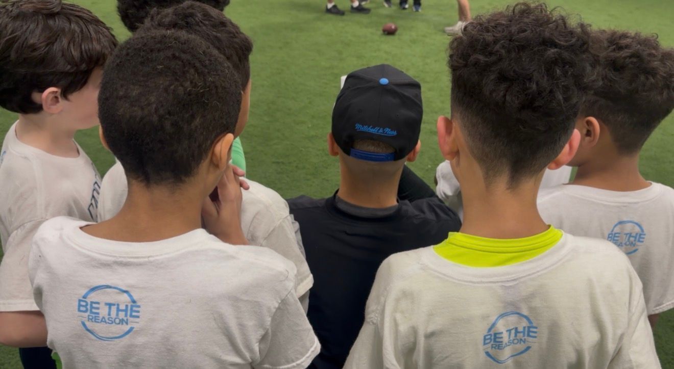 Children in white shirts watch a sports field from the bleachers, seen from behind